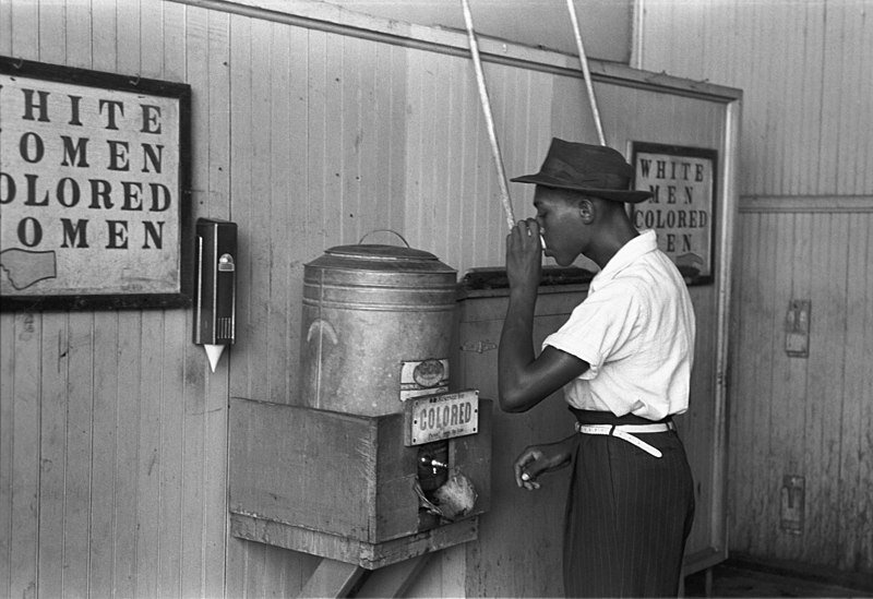 Homme noir de peau buvant à une fontaine devant un panneau instaurant une ségrégation raciale